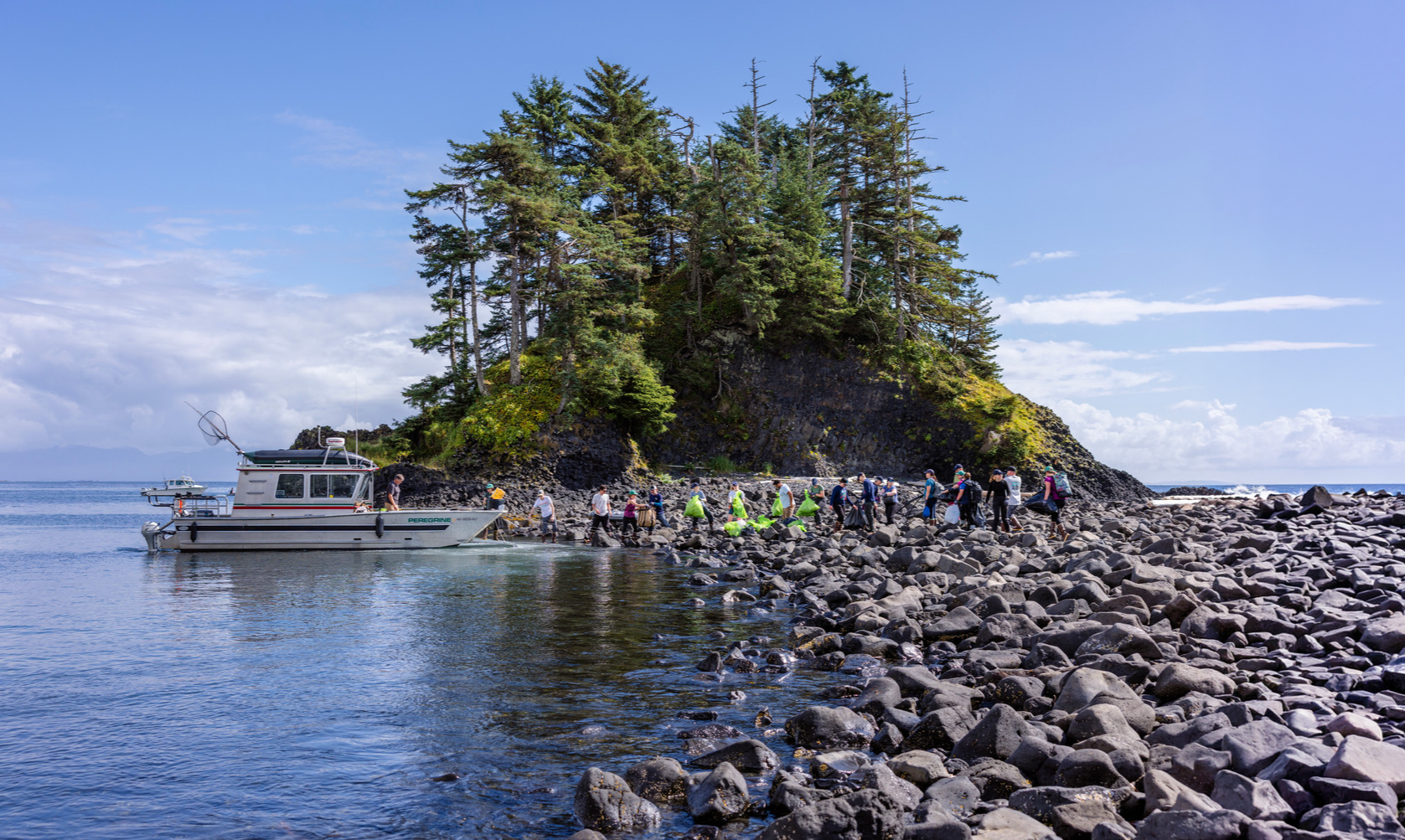 A group of people disembarking a boat onto a rocky shoreline with an island covered in trees in the background.