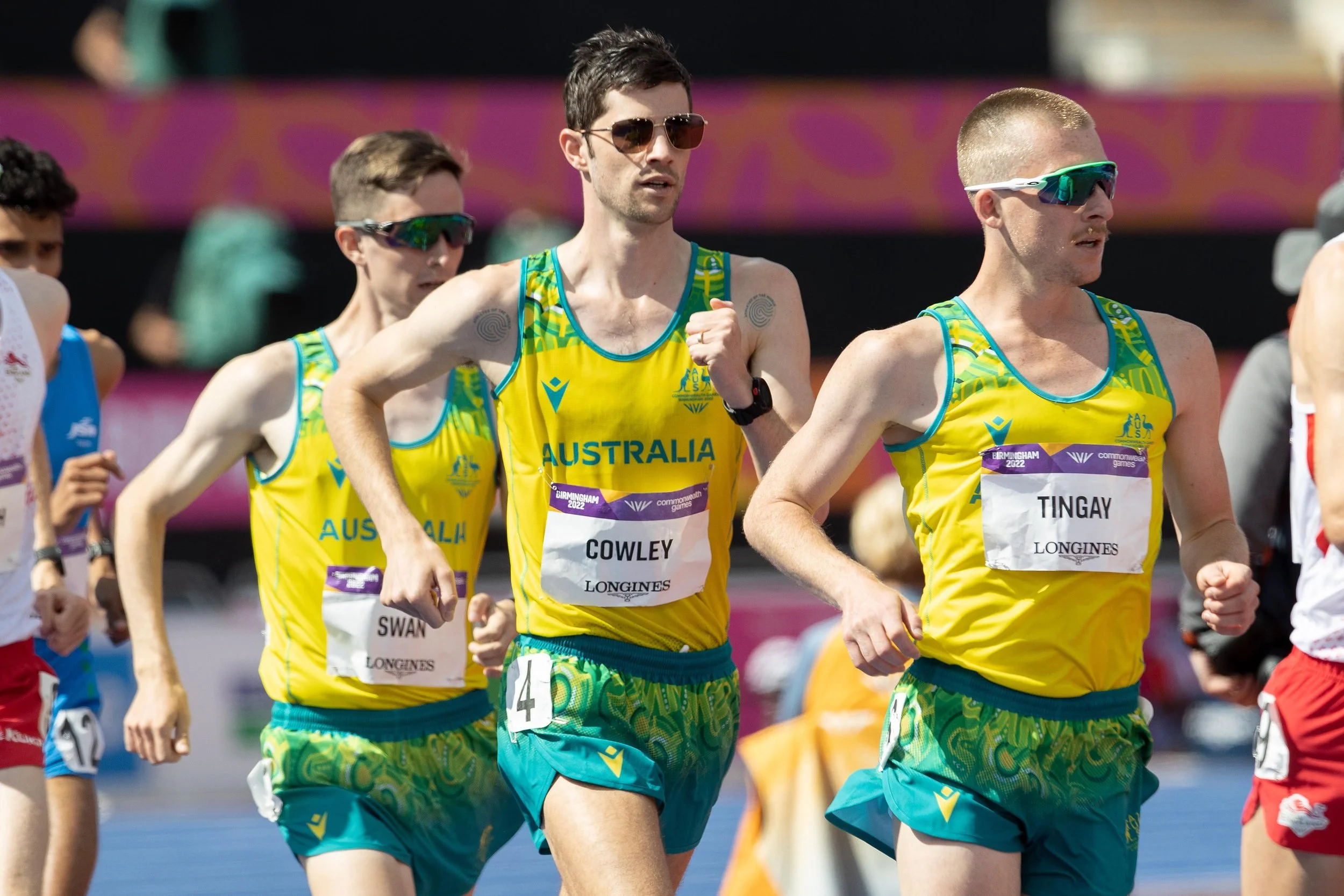 A group of Australian male athletes running in a marathon, wearing yellow and green uniforms with the word 'Australia' on their chests, and sunglasses. They are participating in the Birmingham 2022 Commonwealth Games.