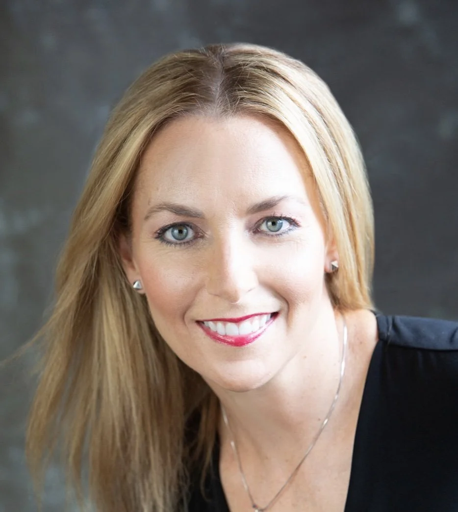 Close-up portrait of a woman with blonde hair, blue eyes, and a bright smile, wearing earrings and a necklace, against a dark background.