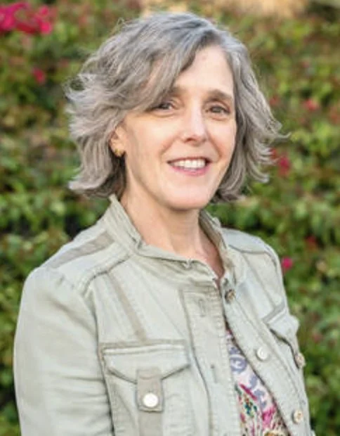 Smiling older woman with gray hair, wearing a light-colored jacket, standing outdoors with greenery and pink flowers in the background.