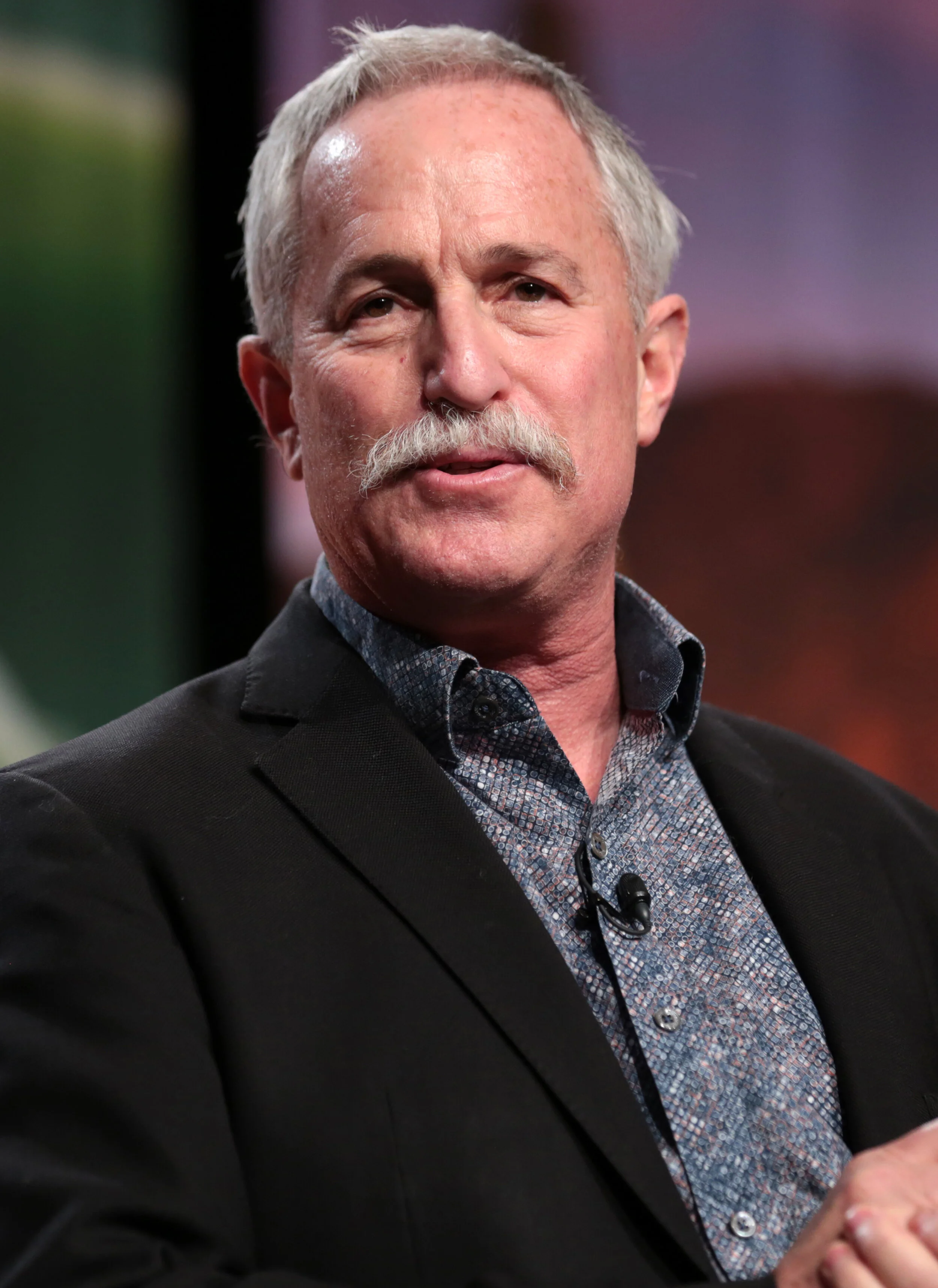 A man with gray hair and a mustache wearing a dark blazer and patterned shirt, speaking or listening in an indoor setting.