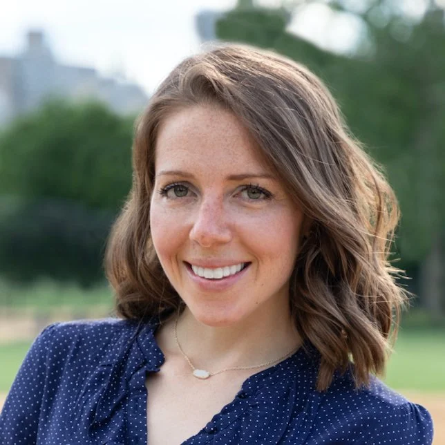 A smiling woman with shoulder-length wavy brown hair, wearing a navy blue top with small white polka dots and a delicate necklace, outdoors with green trees and a blurred cityscape in the background.