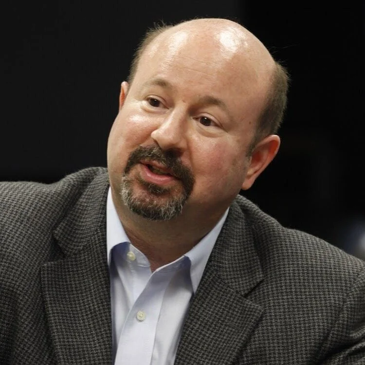 A middle-aged man with a balding head, beard, and mustache, wearing a white dress shirt and a gray, textured blazer, speaking or gesturing during a conversation against a dark background.