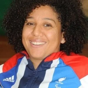 A woman with curly hair smiling, wearing a sports jacket with New Zealand and All Blacks logos.
