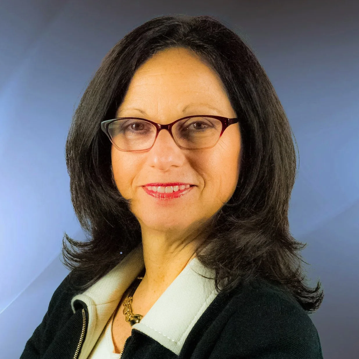 Portrait of a woman with shoulder-length dark hair, wearing glasses, a white collared shirt, a black blazer, and a gold necklace, smiling with a blue gradient background.