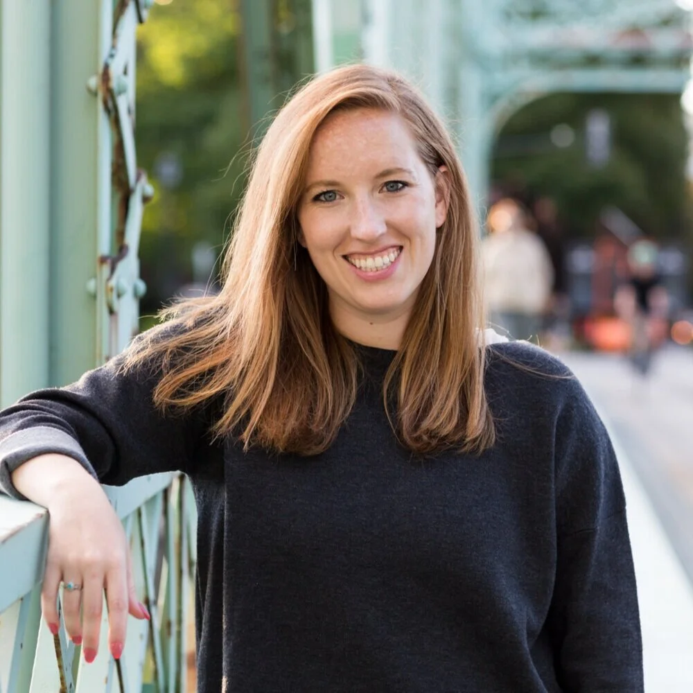A woman with red hair smiling and leaning on a fence outdoors.