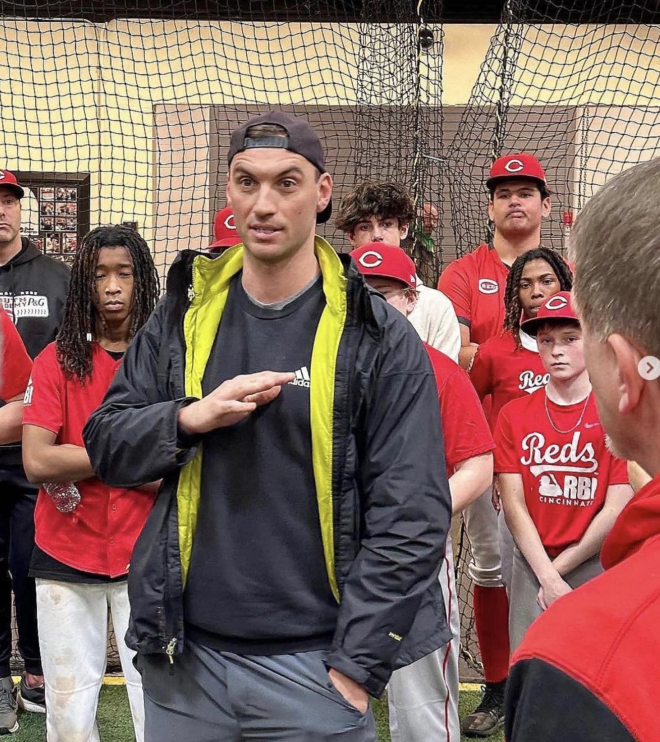 A man in a black cap and jacket is speaking to a group of children and teenagers dressed in Cincinnati Reds and Reds RBI team apparel in an indoor sports facility.