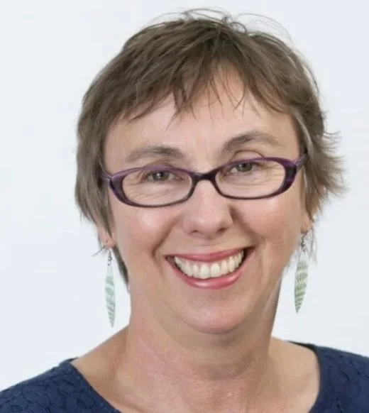 A smiling woman with short brown hair, glasses, and earrings against a plain light background.