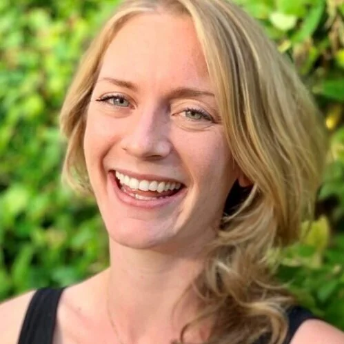 A smiling woman with blonde hair and blue eyes outdoors with green foliage in the background.