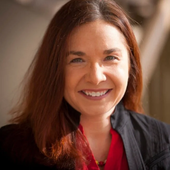 Smiling woman with shoulder-length reddish-brown hair, wearing a black jacket over a red top, indoors with a blurred background.