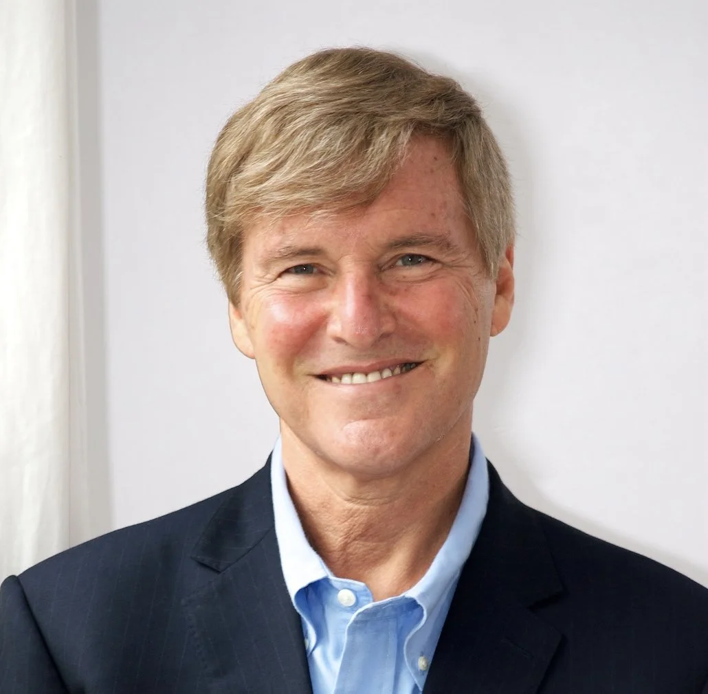 Portrait of a smiling middle-aged man with blond hair, wearing a dark suit jacket and light blue collared shirt, standing in front of a plain light-colored background.