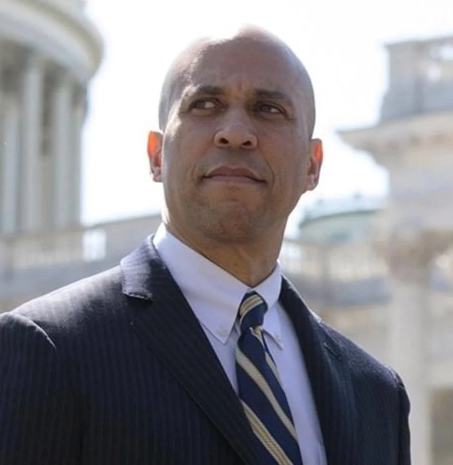 A man in a suit and tie standing outdoors, with a blurred building in the background.