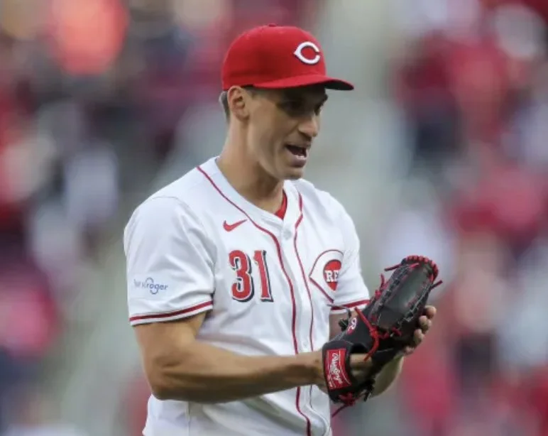 A baseball player wearing a Cincinnati Reds uniform and red cap with a 'C' logo, holding a baseball glove on a baseball field.