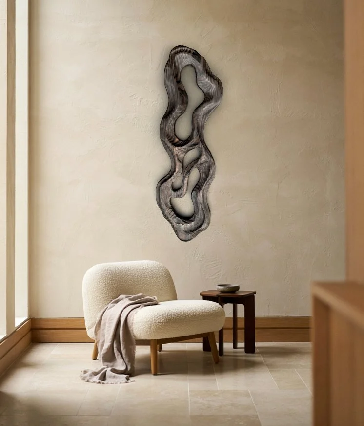 Minimalist interior with a cream-colored plush chair, a beige throw blanket, a small dark wooden side table with a bowl, and abstract wall art with cutouts on a textured cream wall.