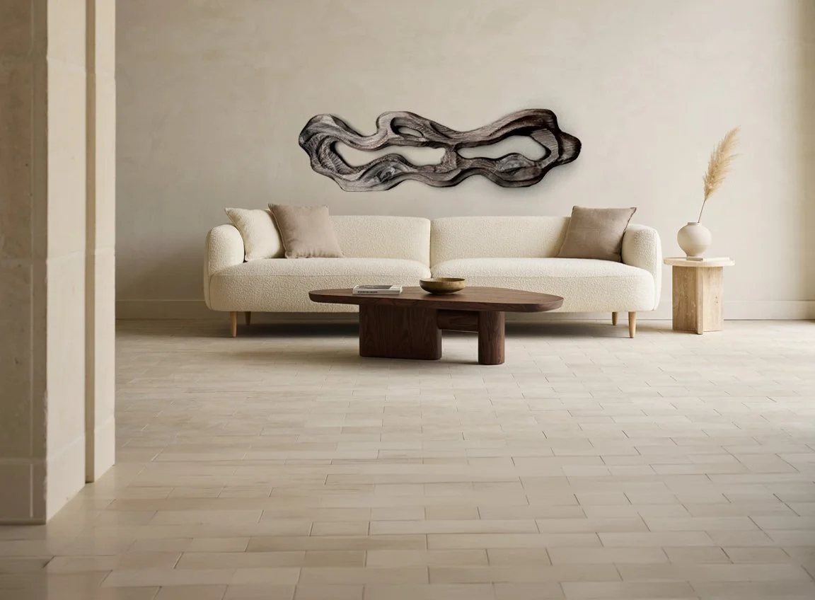 Minimalist living room with a beige sofa, two beige pillows, a wooden coffee table, a side table with a vase and dried plant, and wall art above the sofa.