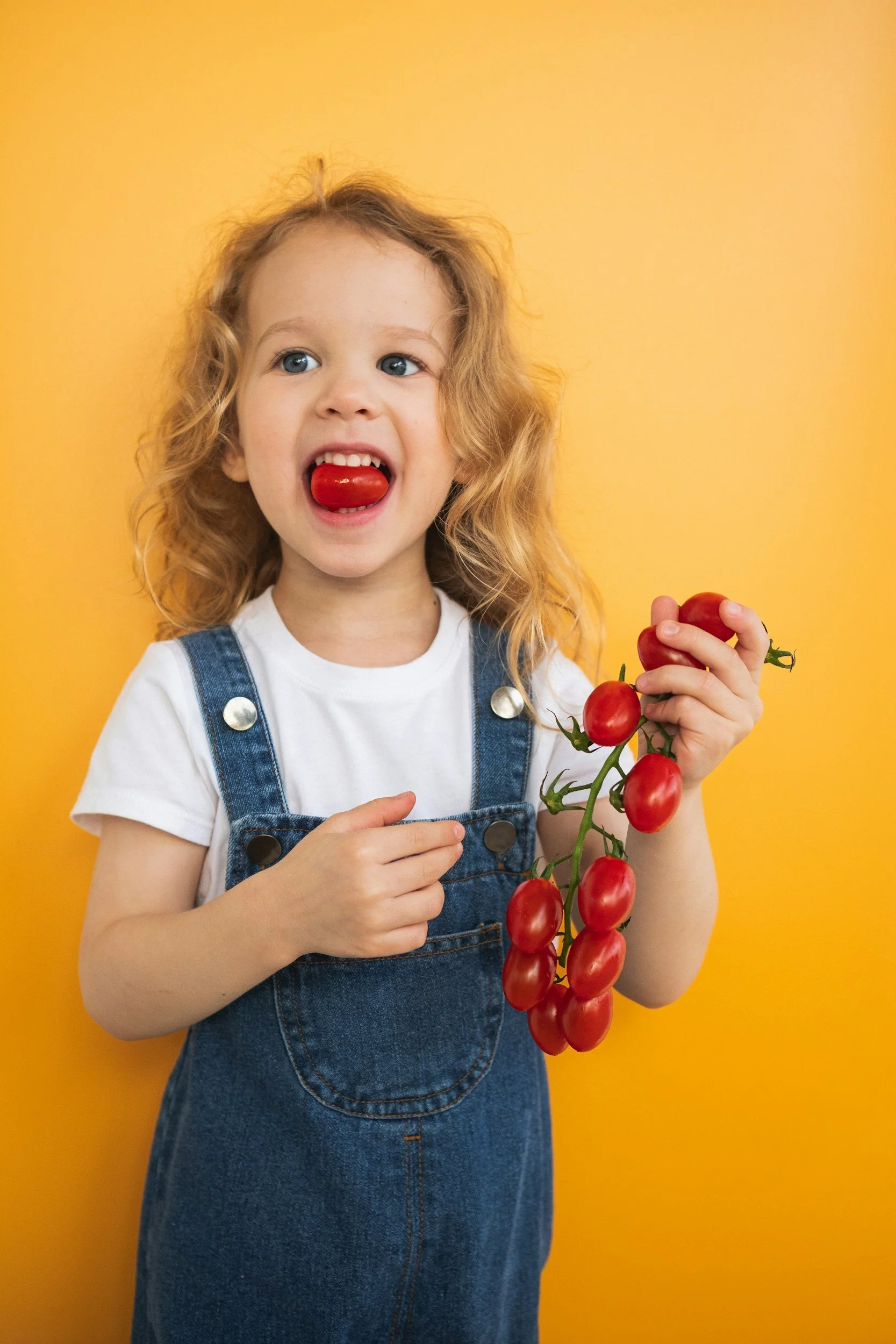 A young girl with curly blonde hair and blue eyes is holding a vine of red cherry tomatoes against a yellow background. She is wearing a white t-shirt and denim overalls, with her tongue out licking a cherry tomato.