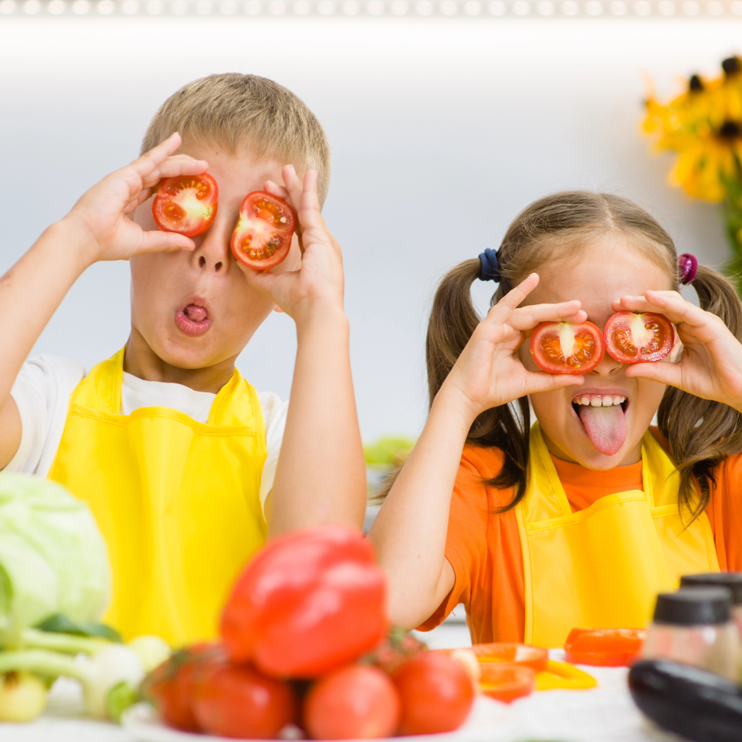 Two children, a boy and a girl, are playing with slices of tomato over their eyes and sticking out their tongues. They are wearing yellow aprons and are at a table with various vegetables, including tomatoes, bell peppers, and lettuce, in front of them.