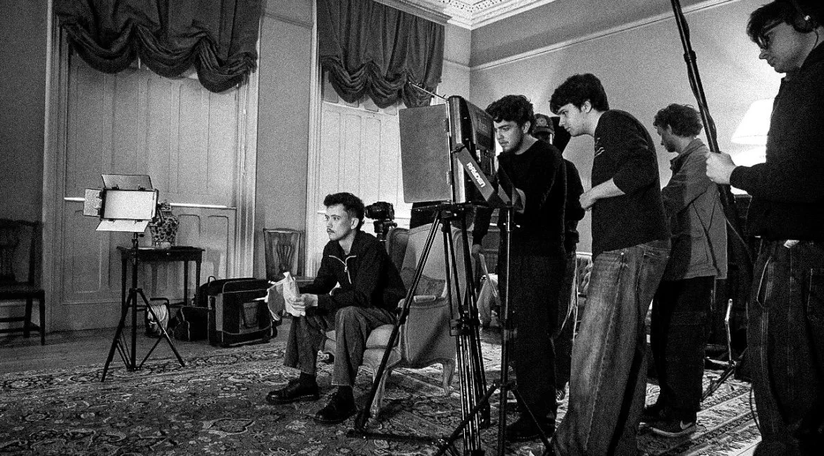 A film crew setting up cameras and lighting in a room with a man seated on a chair holding a paper, ready to film.