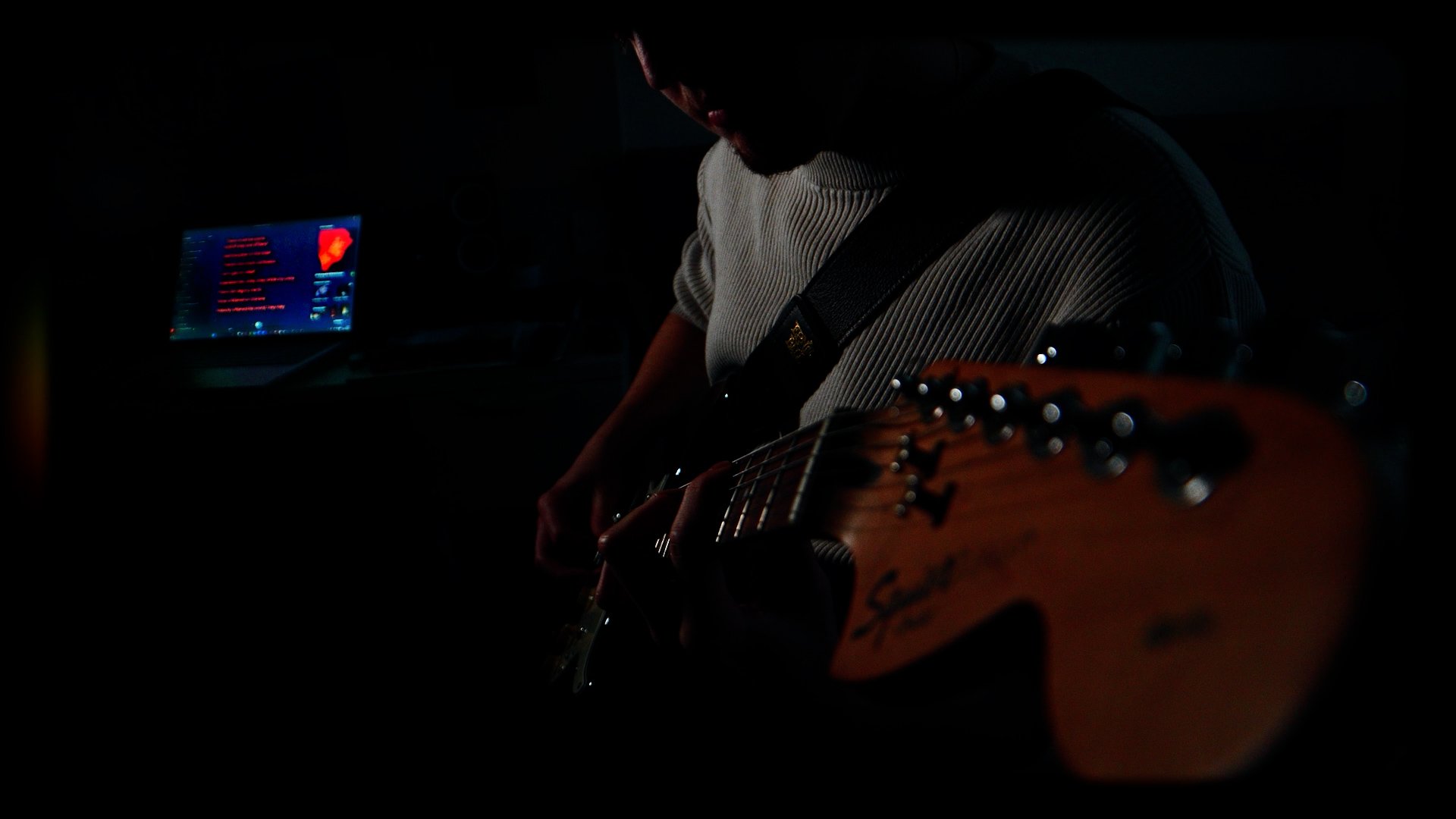A person playing an acoustic guitar in a dark room with a computer monitor displaying a red and blue interface.