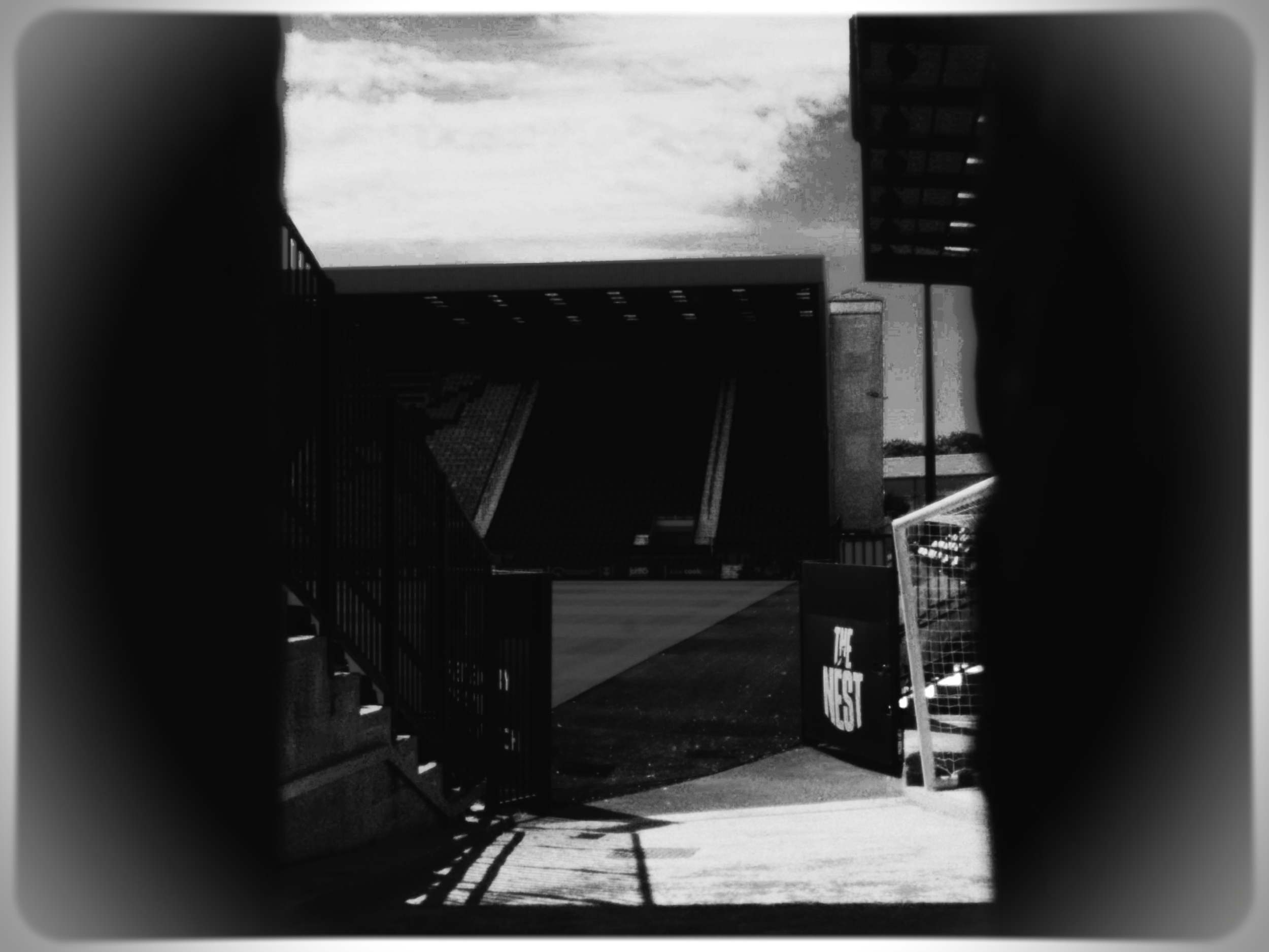 A black and white photograph of an open entrance to a stadium or sports arena, with a metal gate and a sign that reads 'Guest'. The sky is cloudy, and shadows of the gate are cast onto the ground.
