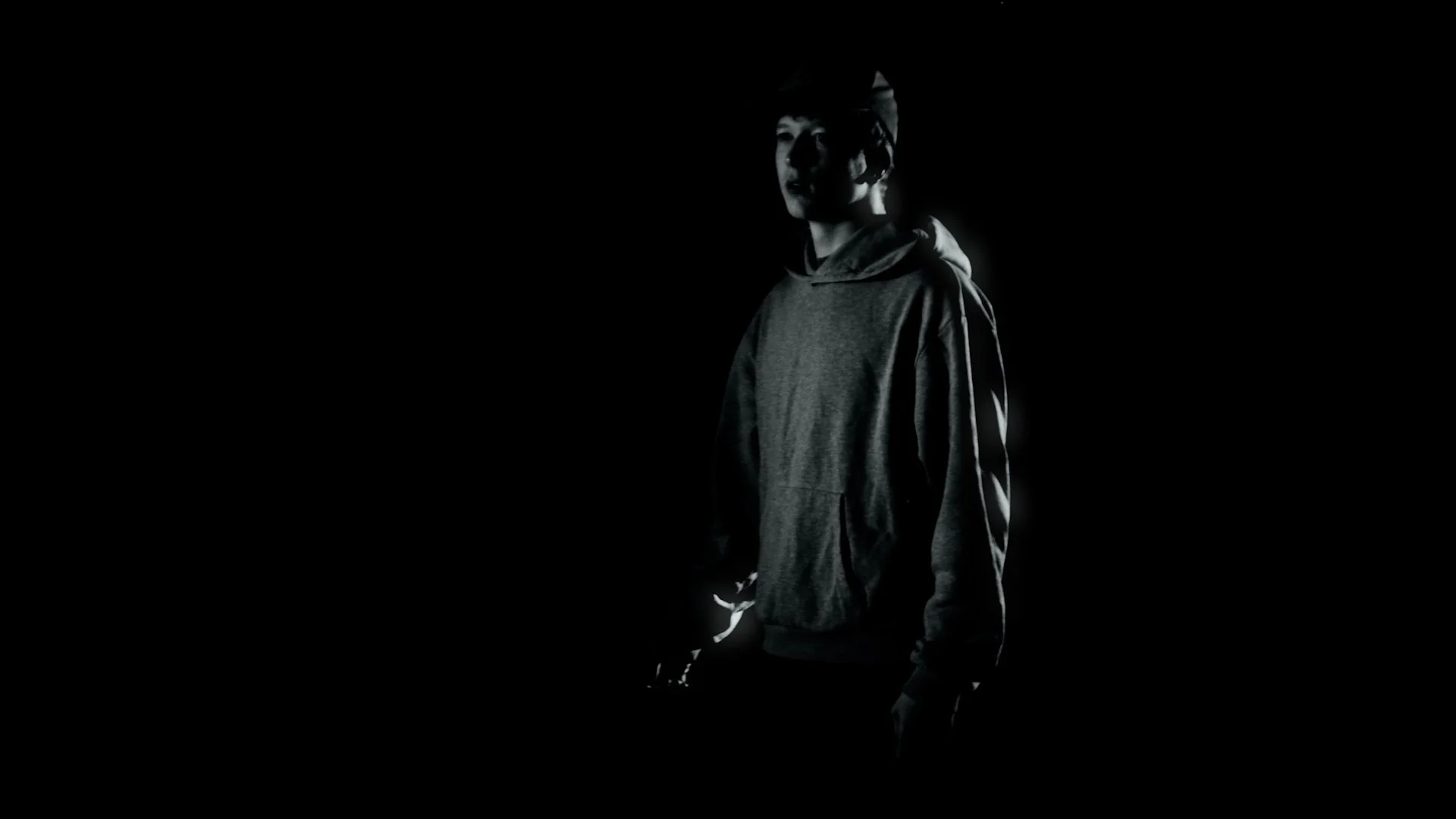 Young man wearing a hoodie and cap standing in a dark environment with minimal lighting highlighting his face and side.