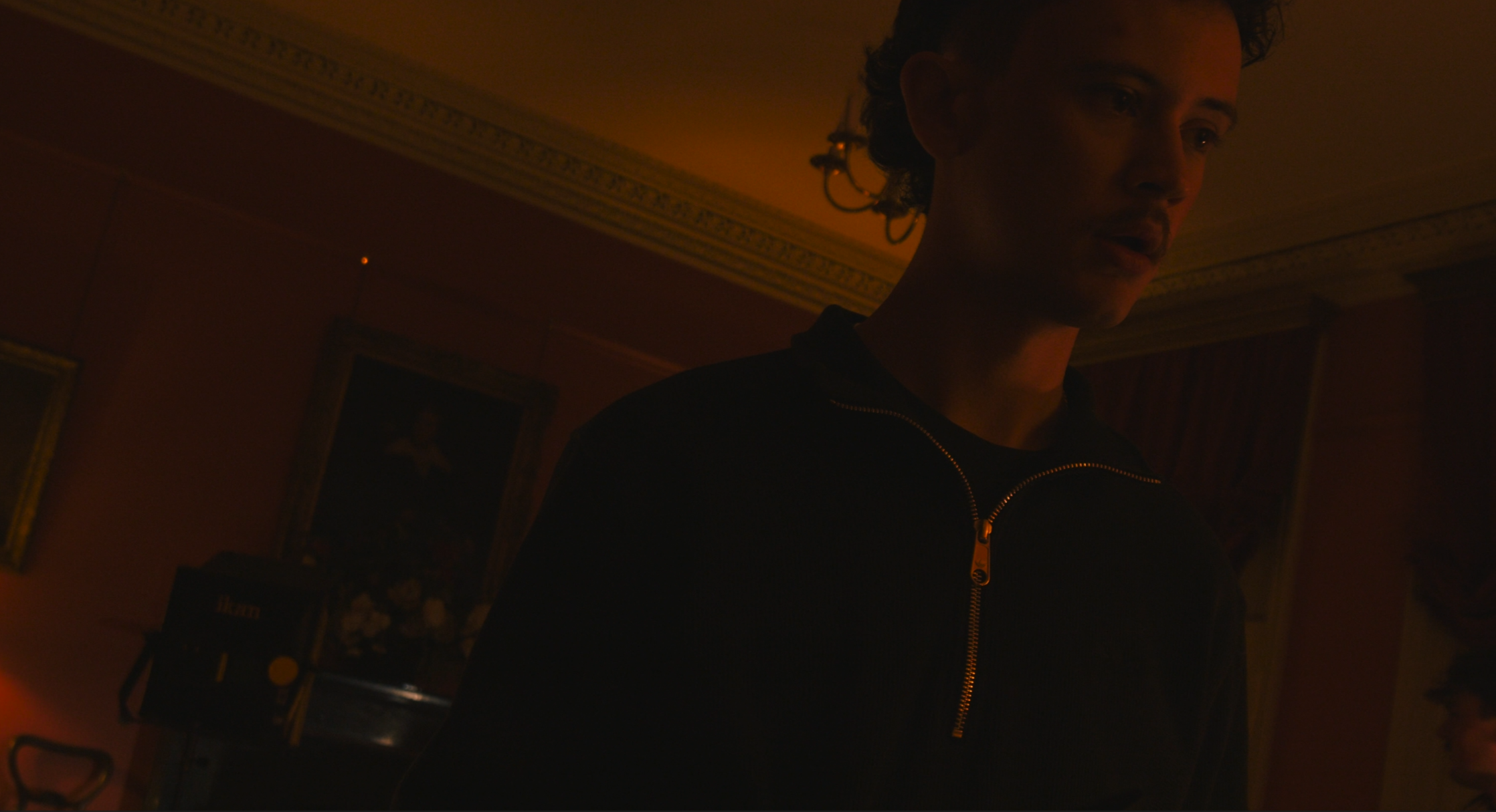 A person with curly hair wearing a black jacket with a zipper, standing in a dimly lit room with ornate moldings and artwork on the walls.