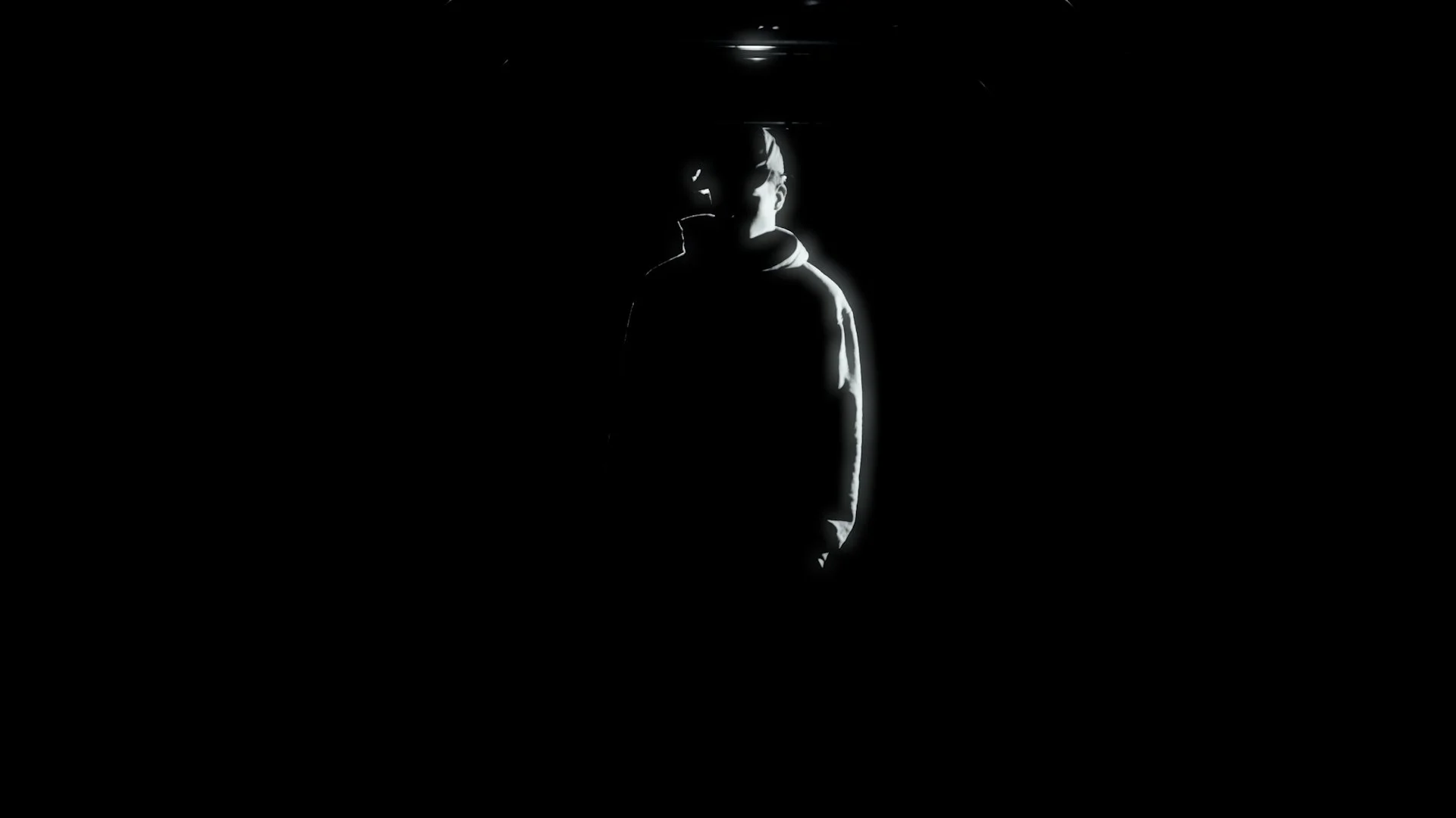 A person standing in darkness with only their outline illuminated, wearing sunglasses and a hoodie.