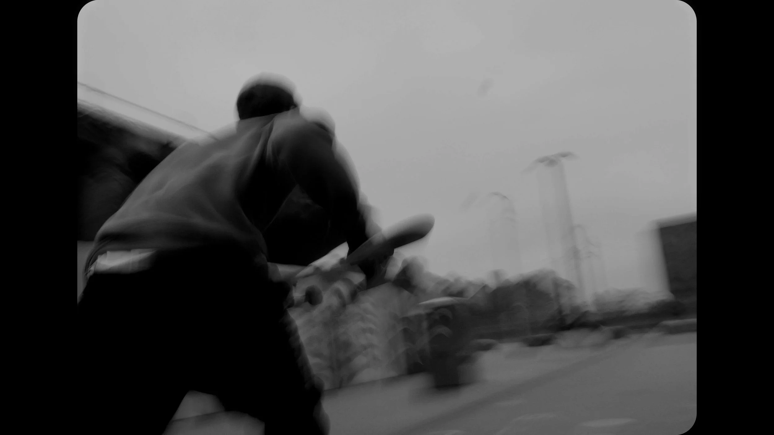 A person riding a motorcycle on the street, seen from behind, in black and white. The sky is cloudy, and there are streetlights and buildings in the background.