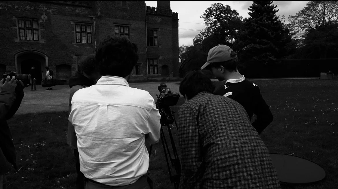 Group of people gathered around a camera outdoors near a historic building, possibly filming or conducting a photography session during the day.