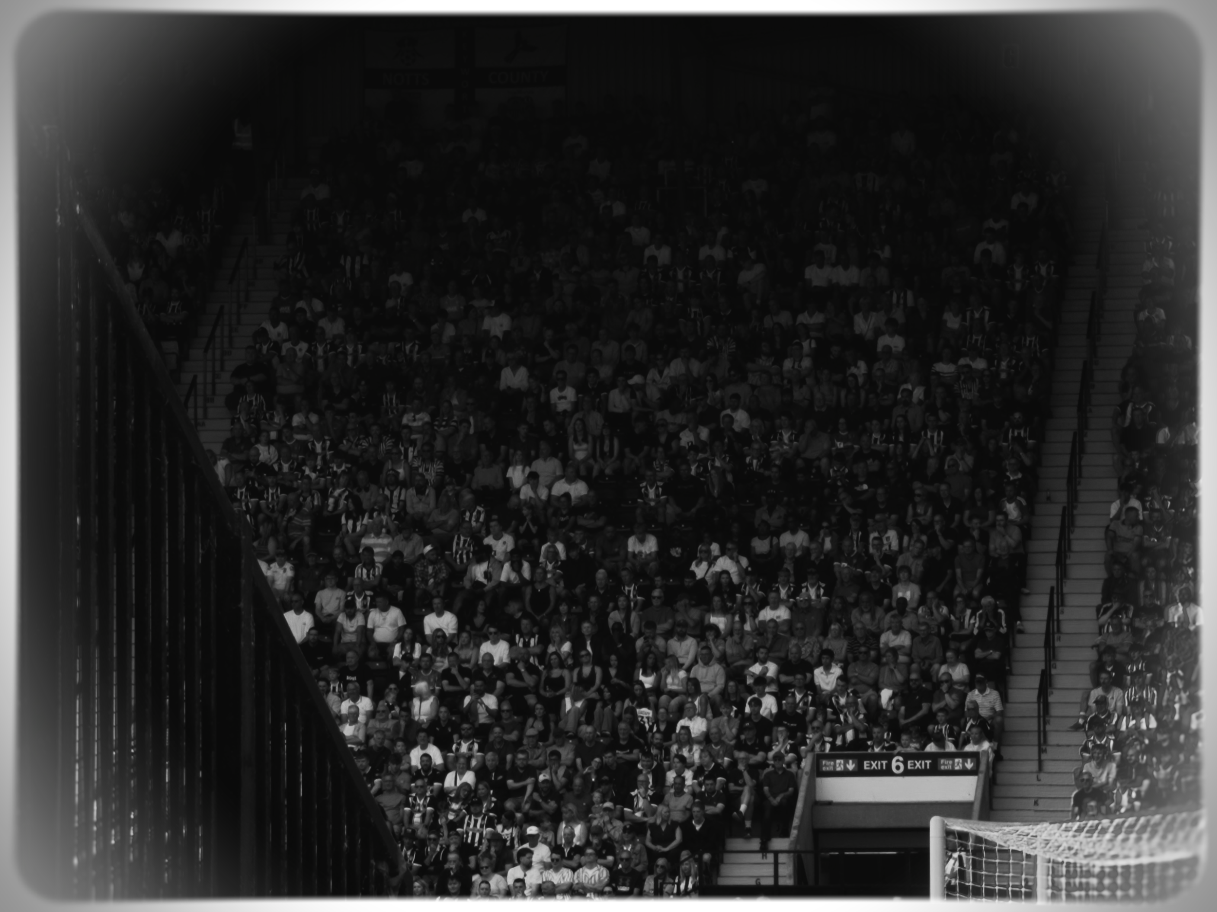 Packed indoor stadium with spectators seated in bleachers, some wearing black and white striped jerseys, possibly at a sports event, with a visible exit sign at the bottom.