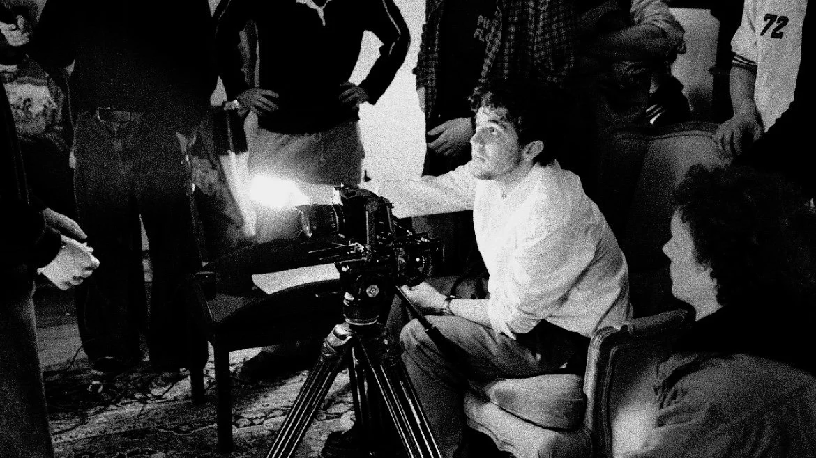 A man operating a camera on a tripod in a room surrounded by multiple people.