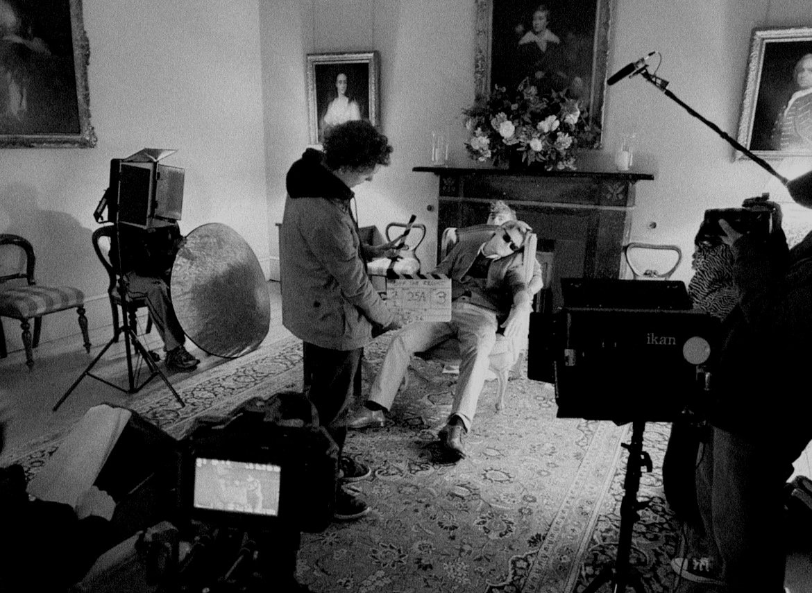 Filming scene with a man sitting on a chair, a woman holding a clapperboard, and crew members operating cameras and equipment in a room with paintings and a fireplace.