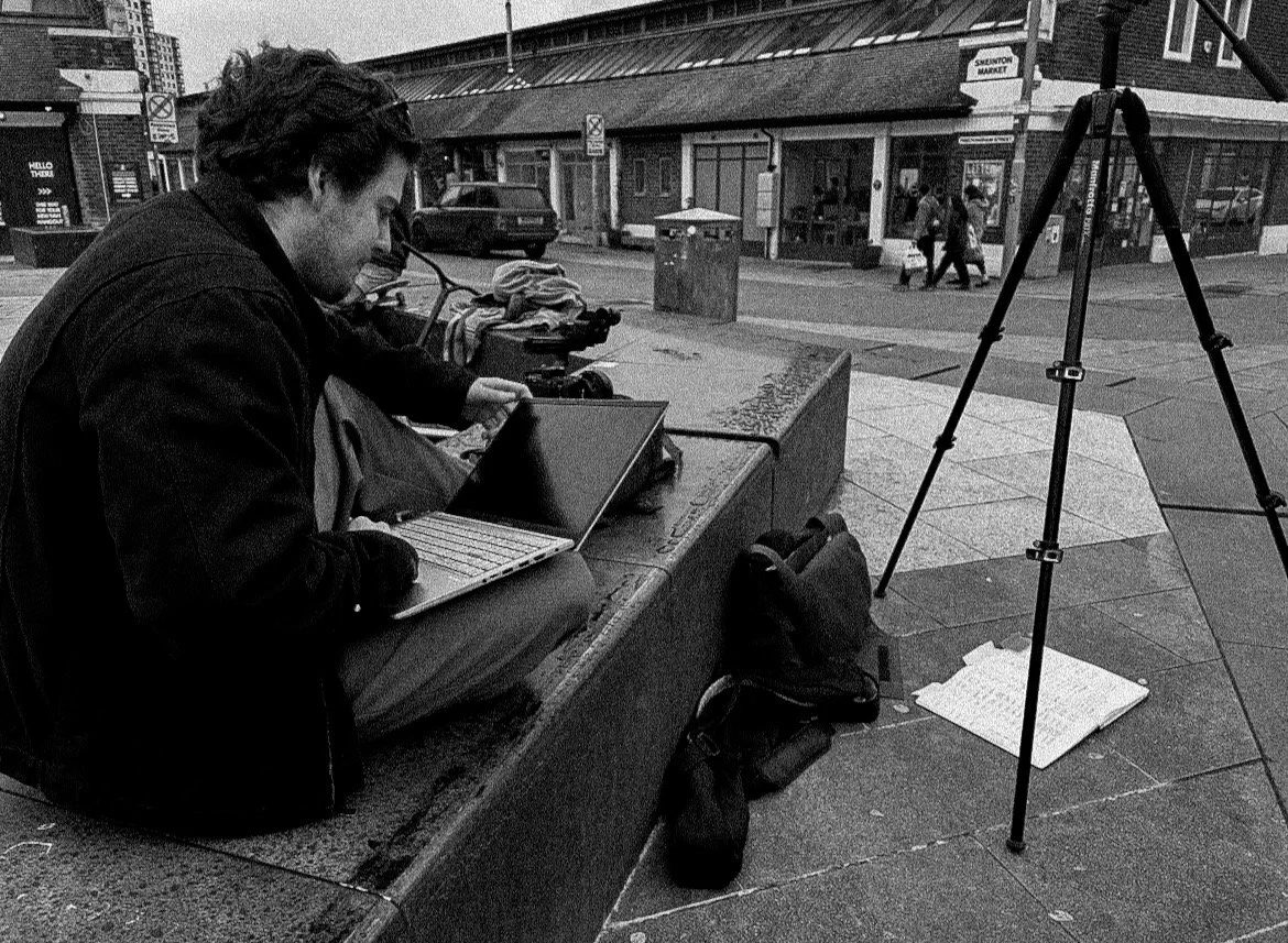 A man sits on a city sidewalk at a bench with a laptop and backpack, working or possibly editing photographs, with a camera on a tripod nearby. In the background, pedestrians are walking on the street and shopfronts are visible.