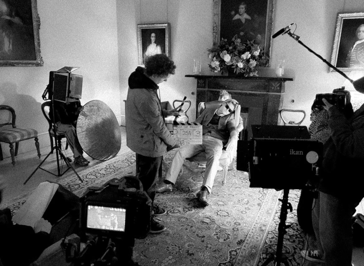 On a film set, a person holds a clapperboard in front of a man lounging in an armchair, surrounded by filming equipment, with portraits hanging on the wall behind.