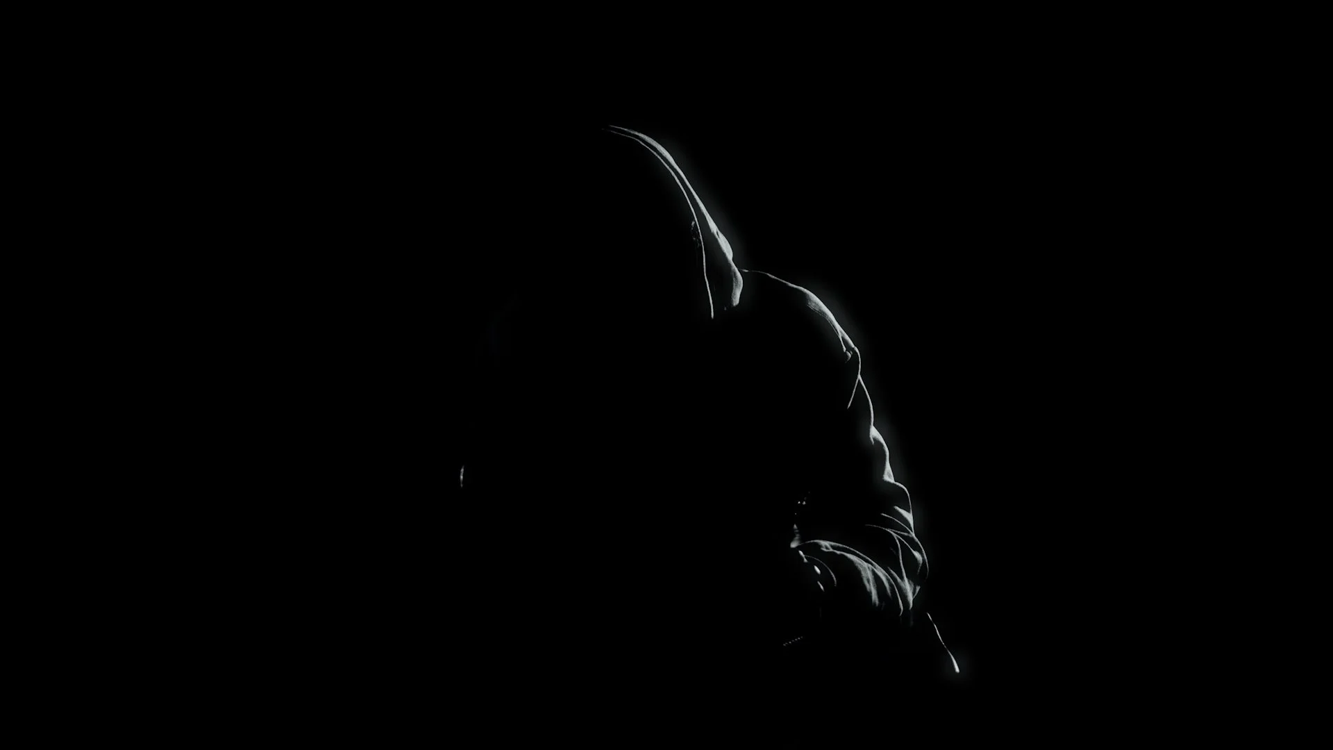 A person wearing a hoodie is partially illuminated in a dark setting, with a focus on the hood and upper body, creating a silhouette effect.