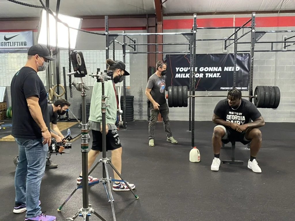 People recording a man in a gym. The man is sitting and lifting a barbell. There are weights, a banner, and gym equipment in the background.