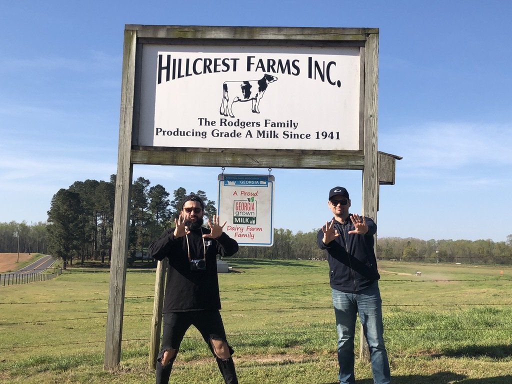 Two men standing in front of a sign at Hillcrest Farms Inc, a dairy farm, with trees and a field in the background, making hand gestures toward the camera.
