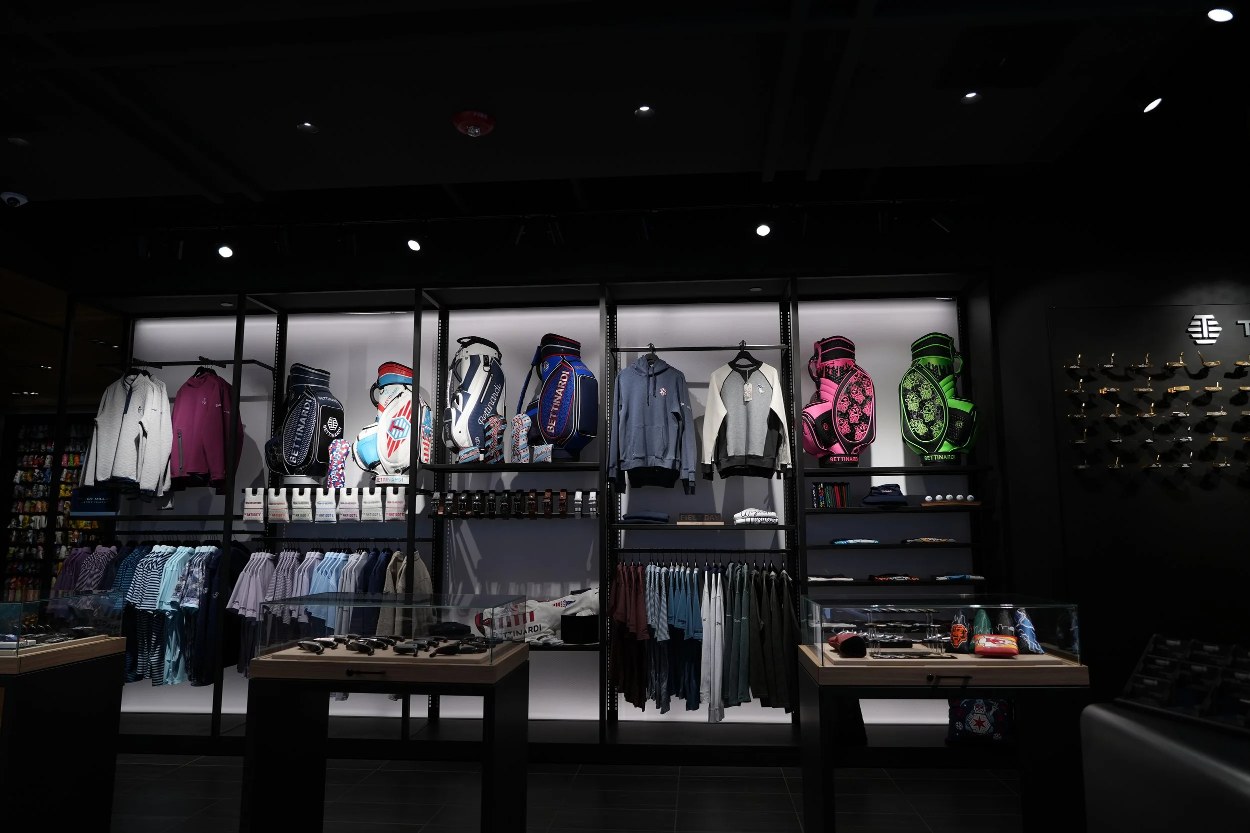 Inside a retail store displaying clothing, golf bags, and accessories on black metal shelves and racks with focused lighting.