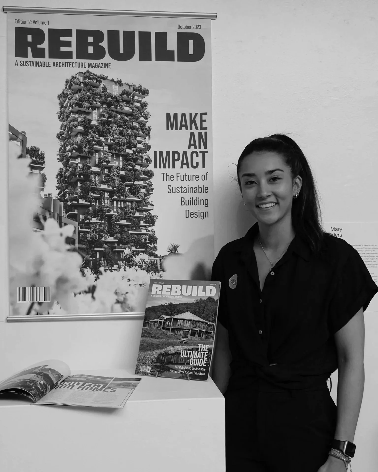 A young woman with long dark hair in a ponytail smiling and standing next to a display of magazines and posters about sustainable architecture and building design.