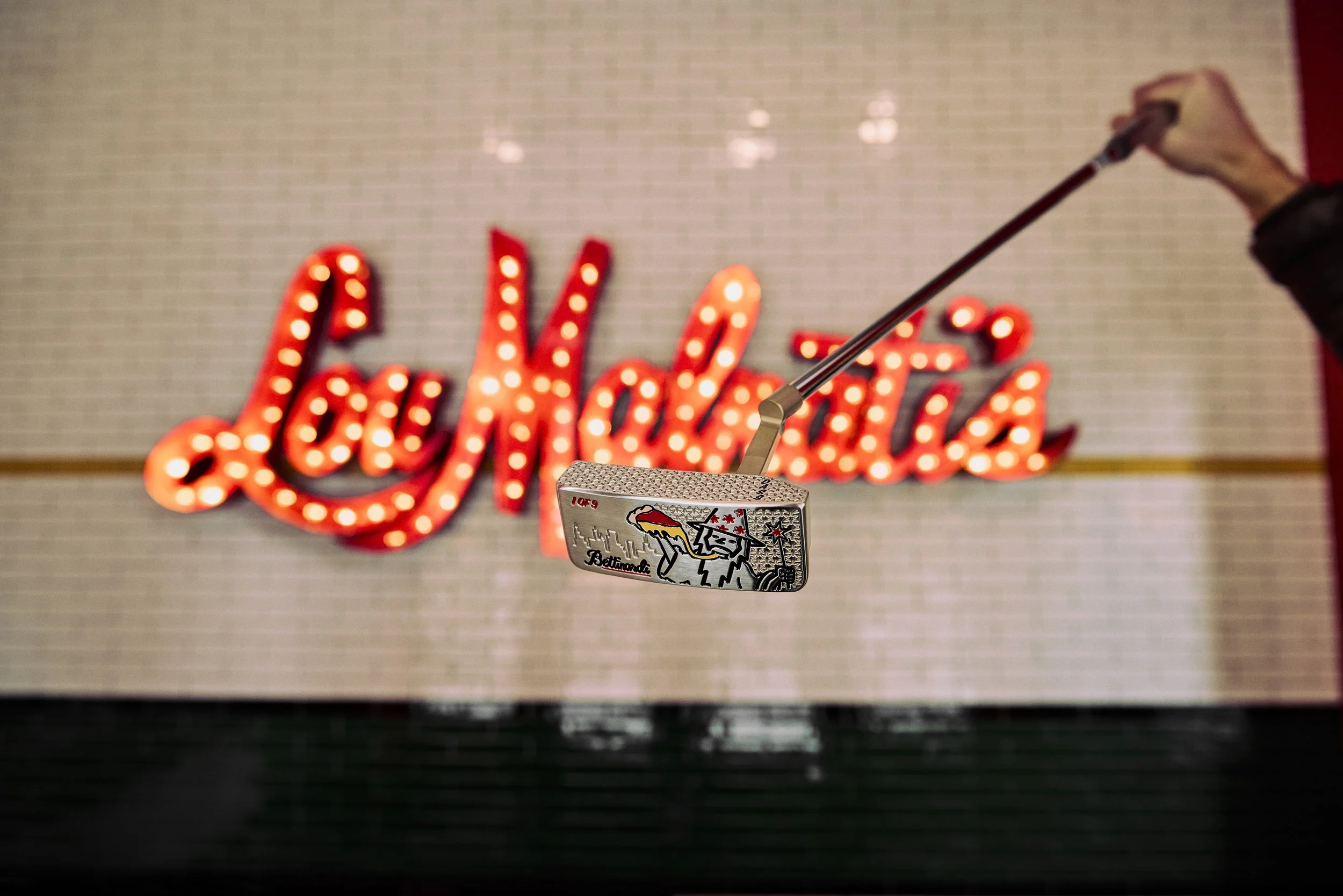 A hand holding a microphone is pointed at a small silver lighter with a graphic of a skeleton playing the guitar and the words 'Betinotal' on it, in front of a red illuminated sign that spells 'cookies' in cursive with light bulbs.