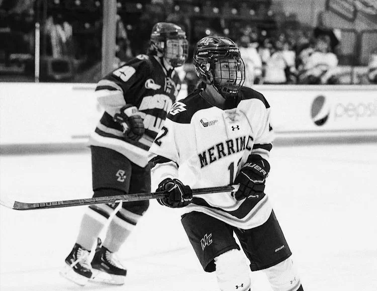 Two hockey players on the ice during a game, one in a white jersey with 'MERRIMACK' written on it, and the other in a darker jersey, both wearing helmets and hockey gear.