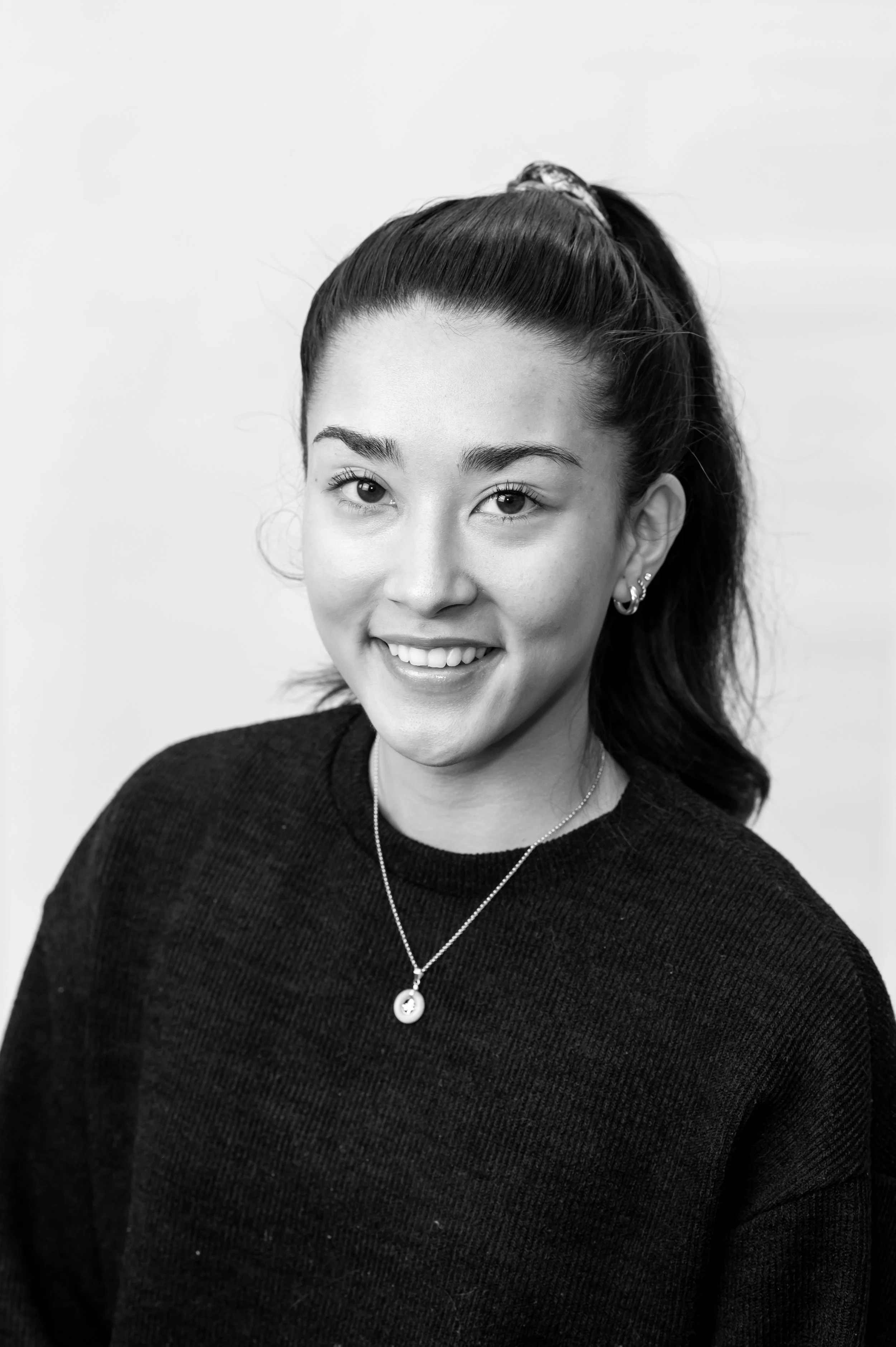 Black and white portrait of a woman with dark hair in a ponytail, smiling, wearing earrings, a necklace with a circular pendant, and a dark sweater.