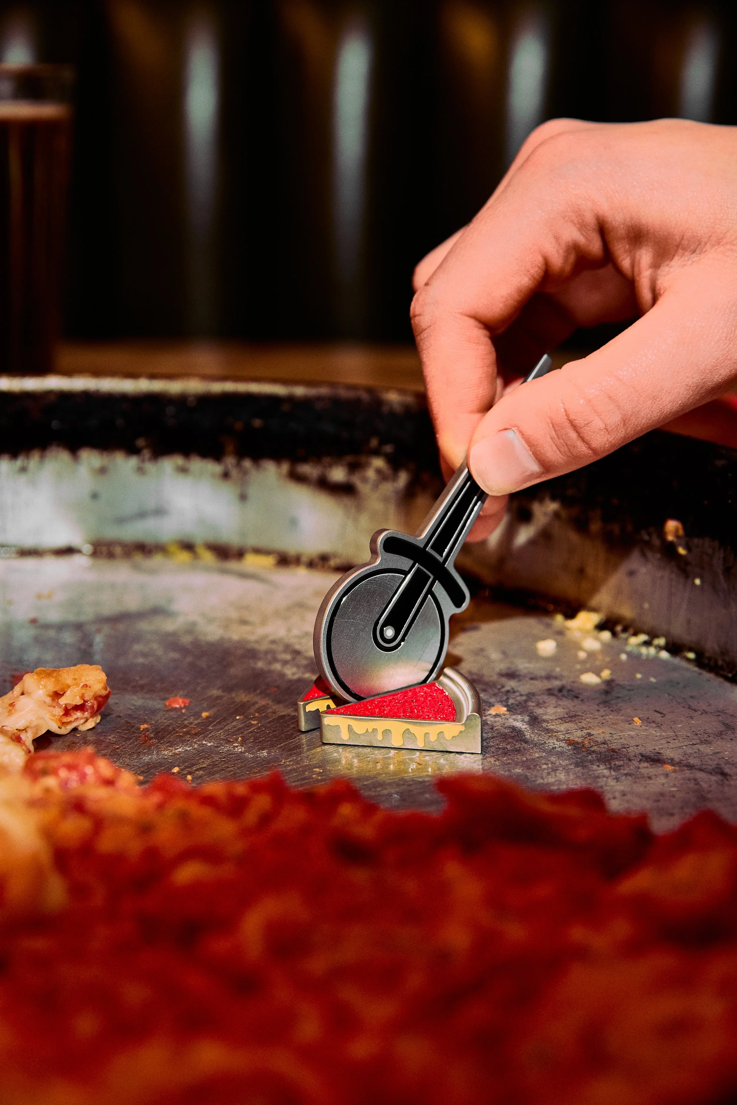A person's hand holding a pizza cutter and slicing through pizza on a baking sheet, with a blurred background of a kitchen or restaurant.