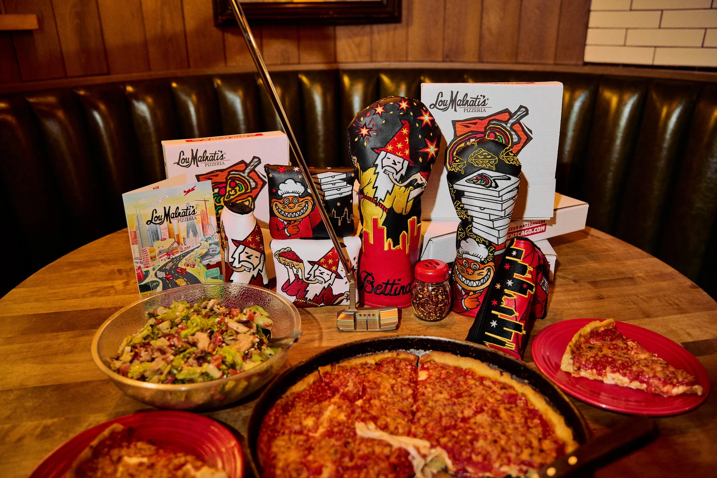 A table with two pizzas, a salad in a glass bowl, and several decorative items including oven mitts and boxes from Lou Malnati's Pizzeria.