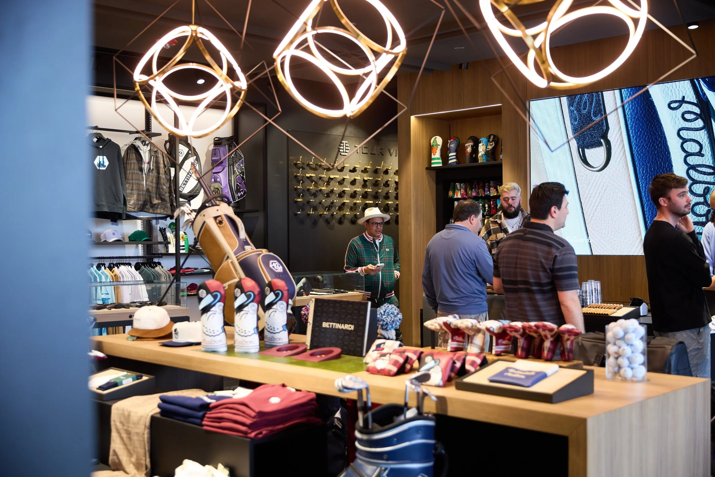 Interior of a retail store with clothing, golf equipment, and accessories on display, customers shopping, and modern lighting fixtures overhead.