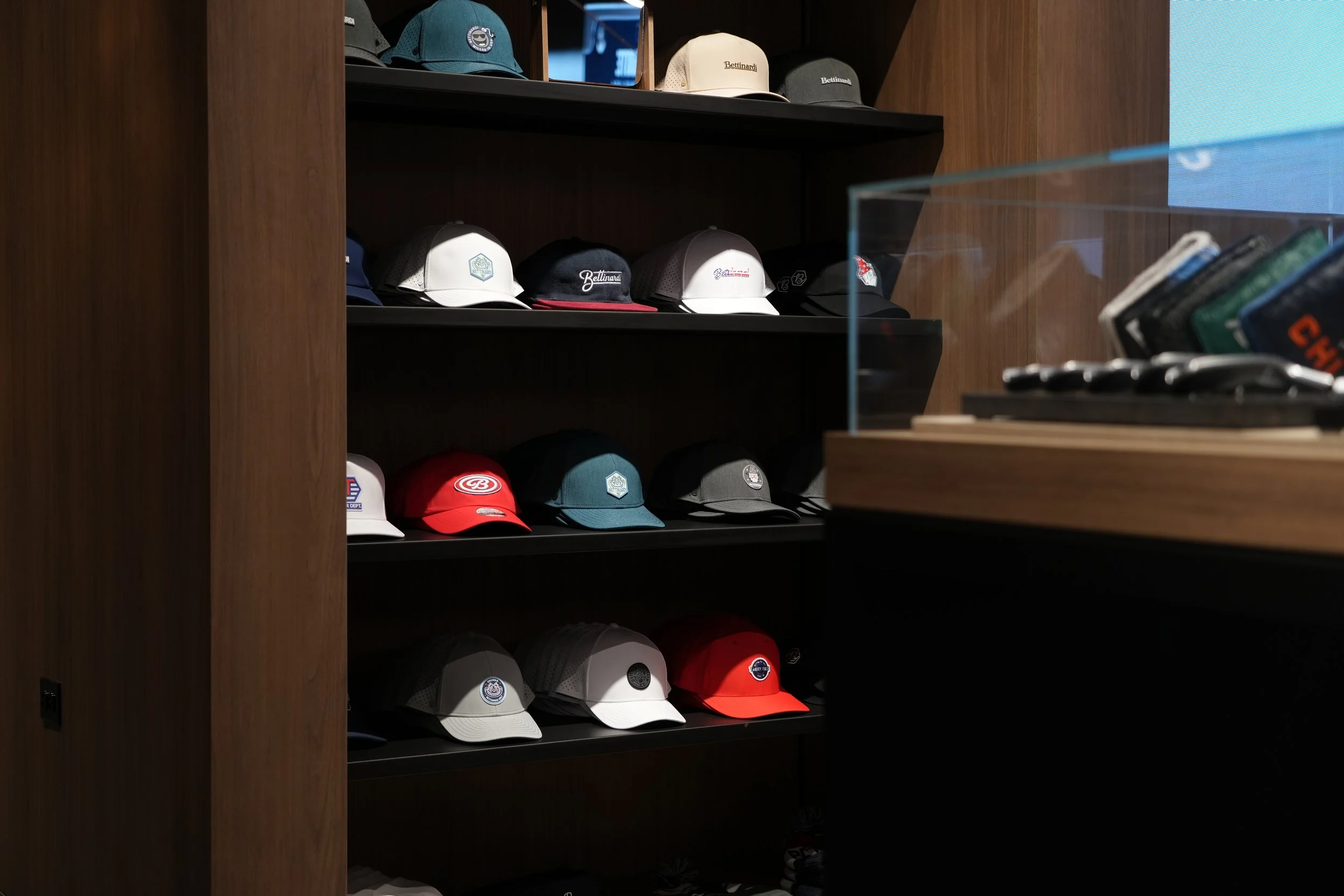A display shelf with various baseball caps, including white, black, red, and teal, many with logos or text, and a glass protector on the right side.