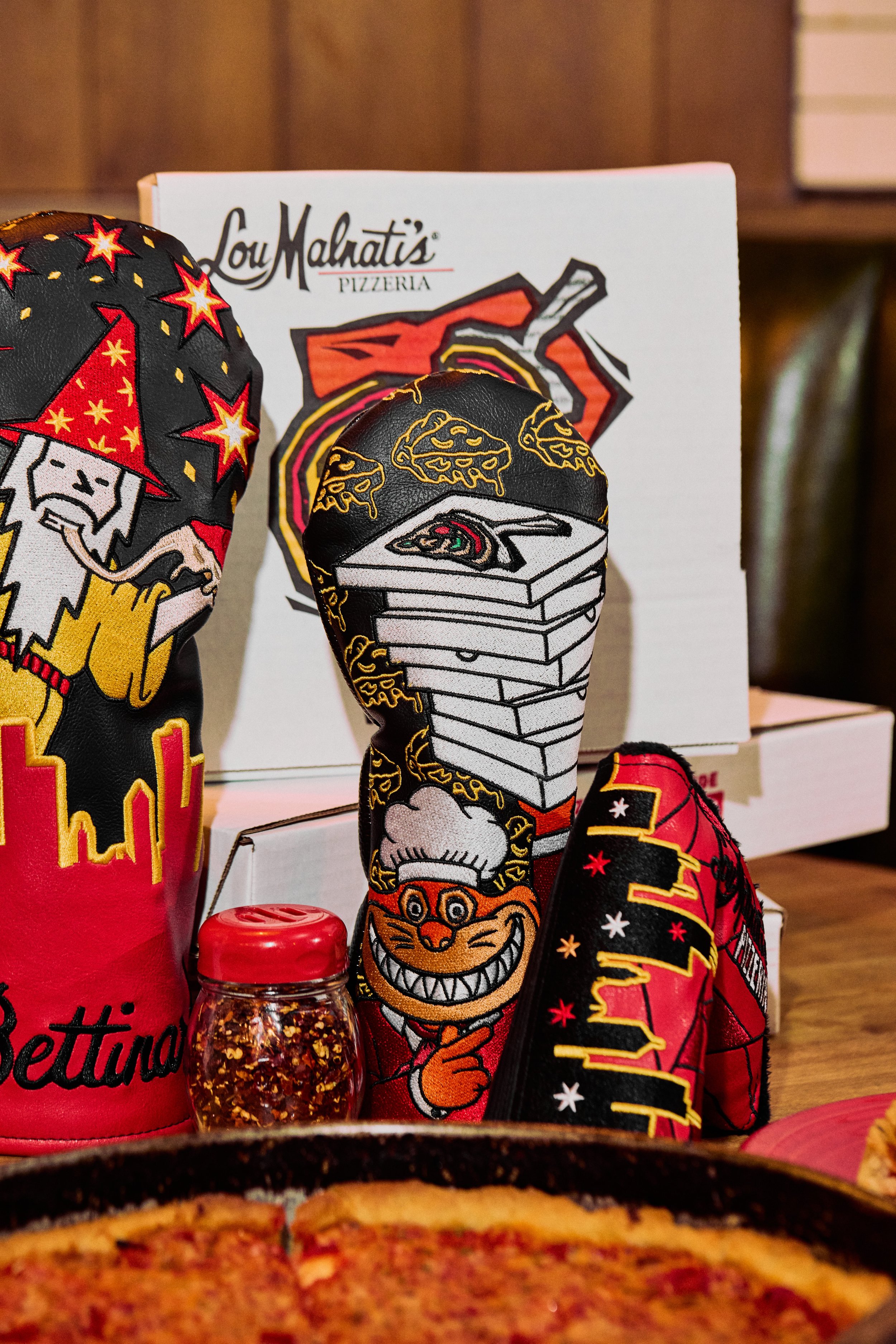 Display of colorful oven mitts with cartoon characters and graphics, a bottle of chili flakes, and a pizza in the foreground at Lou Malnati's Pizzeria.