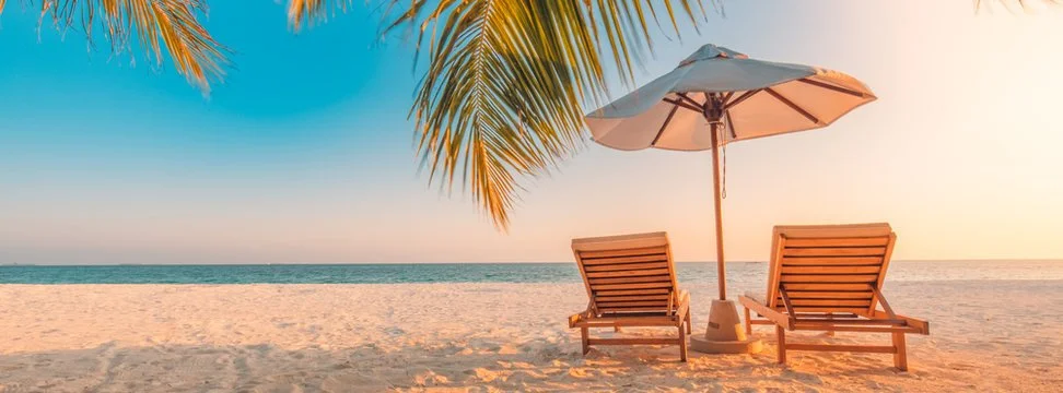 Two wooden lounge chairs and a large beach umbrella on sandy beach with palm leaves overhead, ocean in the background during sunset or sunrise.