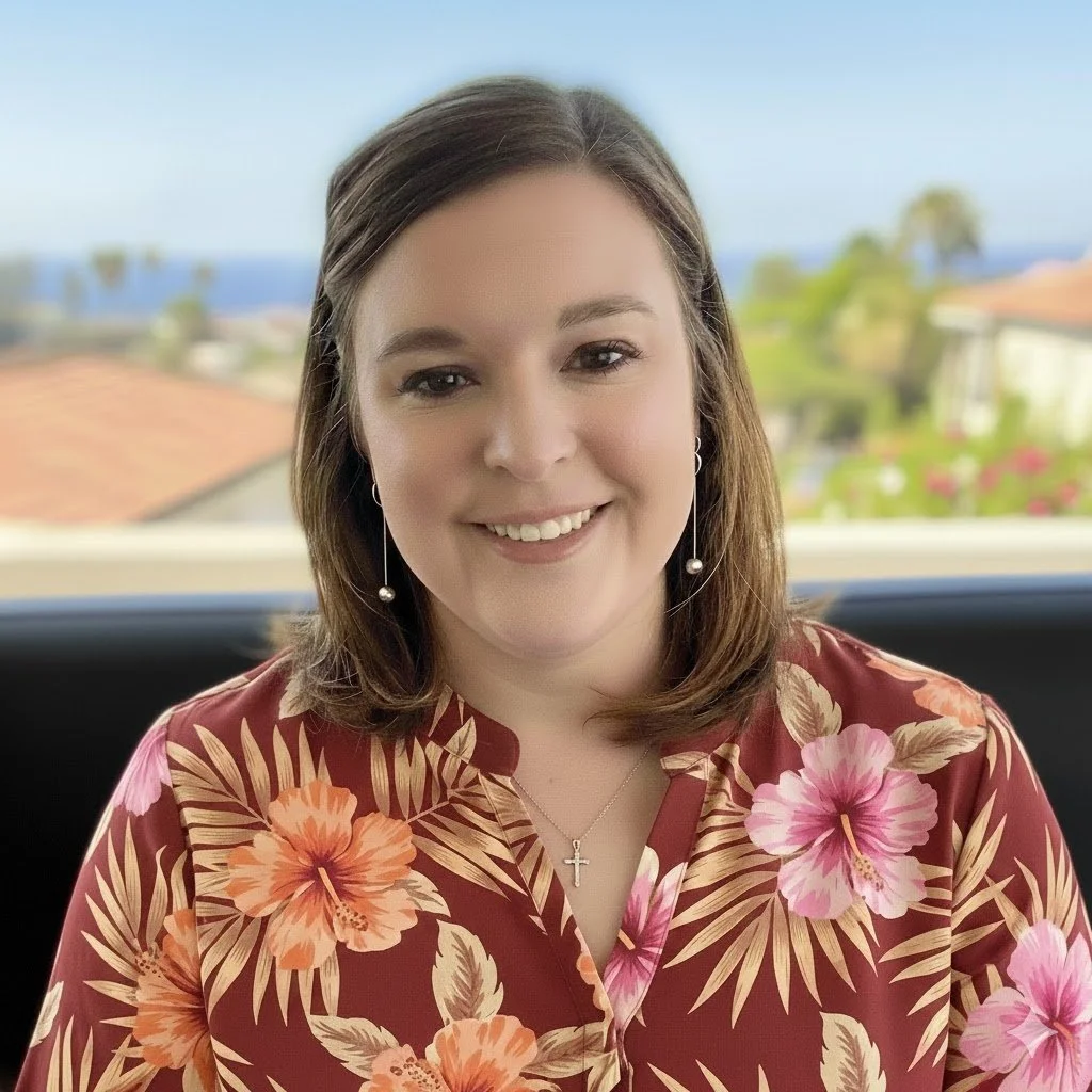 A smiling woman with shoulder-length brown hair wearing a red floral shirt, earrings, and a cross necklace, sitting outdoors with a blurred background of houses, trees, and a blue sky.