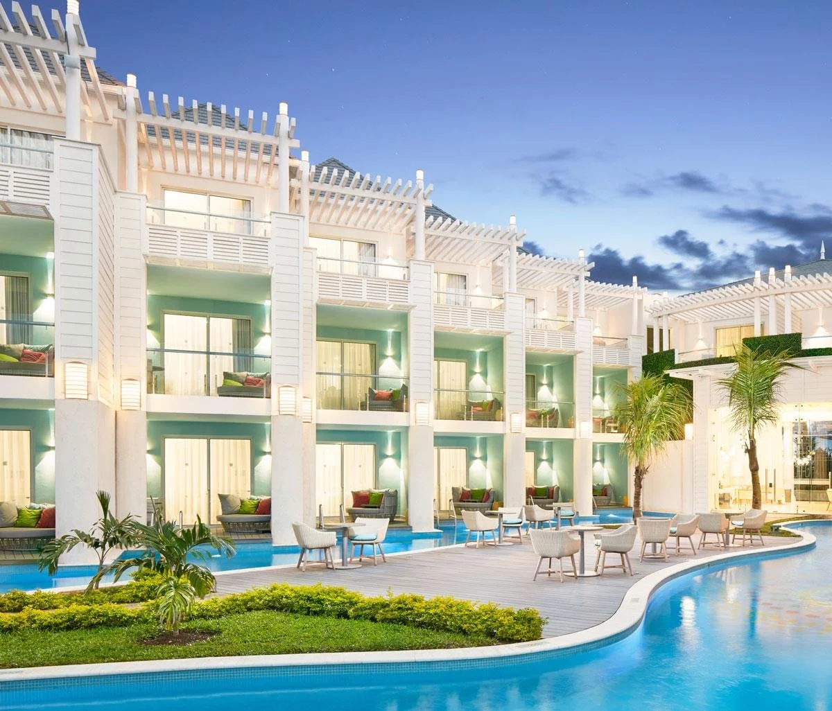 Night view of a modern hotel with a swimming pool, outdoor seating, palm trees, and balconies, illuminated with soft lighting.